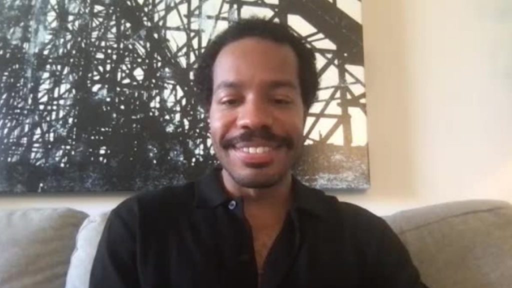 A man with short curly hair and a mustache sits on a couch, smiling at the camera. There is abstract black and white wall art behind him.