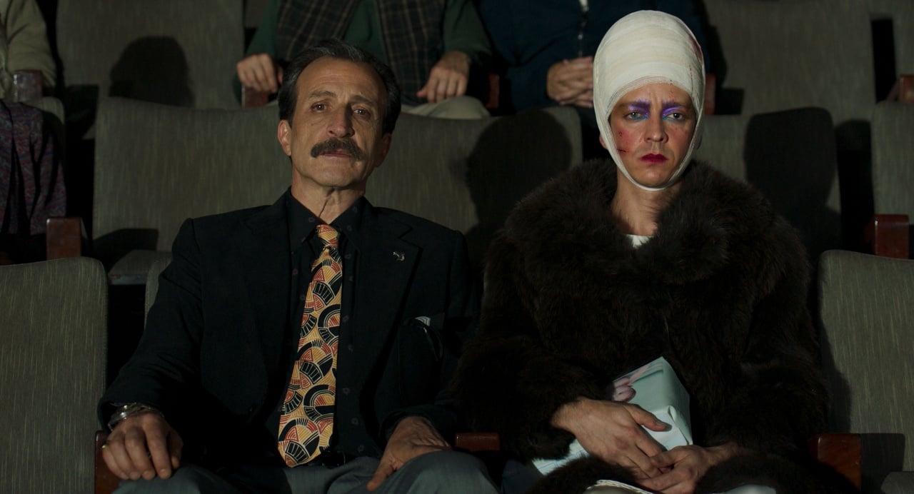 A man in a suit and a woman with a bandaged head, heavy makeup, and a fur coat sit side by side in an empty theater.
