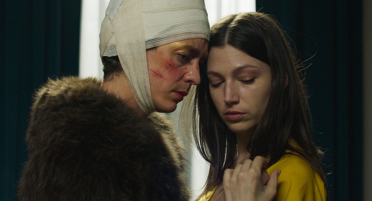 A person with a bandaged head and facial bruises stands close to a woman in a yellow top, both appearing serious and introspective.