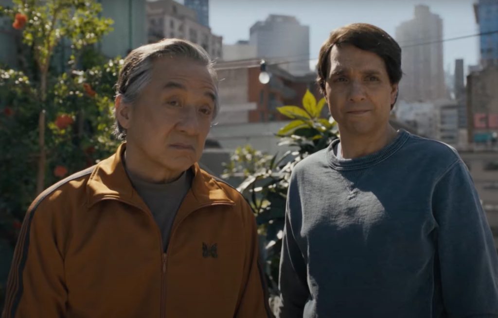 Two men stand outdoors in a city garden, one wearing an orange jacket and the other in a blue sweatshirt, with buildings visible in the background.