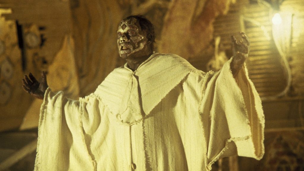 A person wearing a white robe stands with arms raised; their face and head are covered in dark makeup or paint, with dramatic lighting and a blurred background.