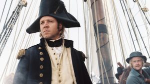 A man in a navy officer’s uniform stands on the deck of a sailing ship, with ropes and masts visible in the background and another crew member looking on.