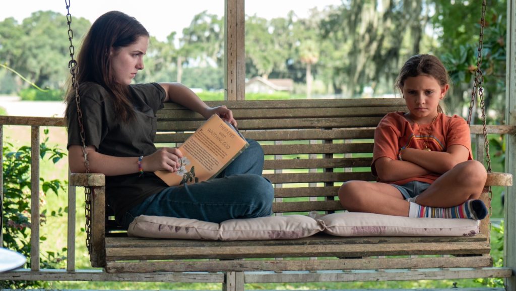 Two girls sit on a porch swing; one is holding an open book and looking at the other, who sits cross-legged with arms folded, appearing upset.