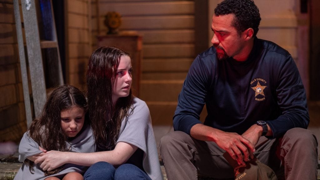A sheriff sits beside two distressed girls wrapped in blankets on a porch at night, with one girl comforting the other.