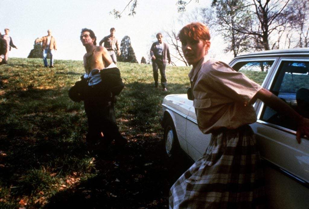 A group of people walk on a grassy hill near a white car; one person in front holds the car door while others approach in the background.