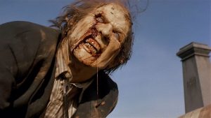 A person with decayed skin, blood around the mouth, and torn clothing appears as a zombie, leaning forward outdoors near a gravestone.