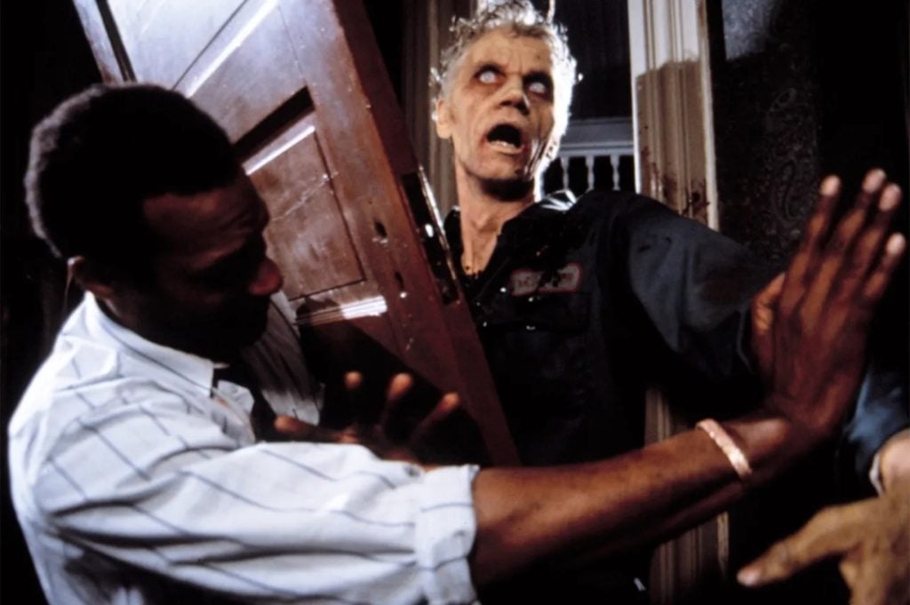A man pushes against a wooden door as a zombie with pale skin and white eyes tries to break through from the other side.