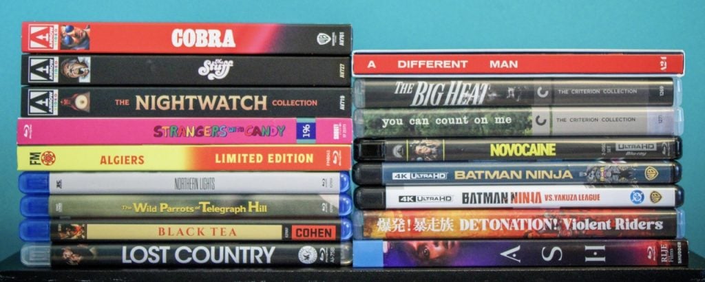 A collection of DVDs and Blu-rays, stacked in two columns, featuring various movie and TV titles with colorful spines against a turquoise background.