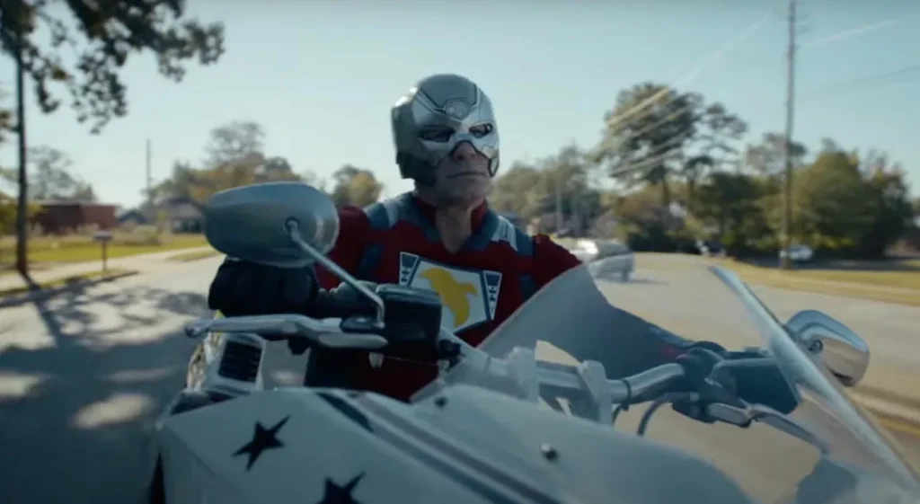 A person in a superhero costume and helmet rides a white motorcycle with star decorations on a suburban street.