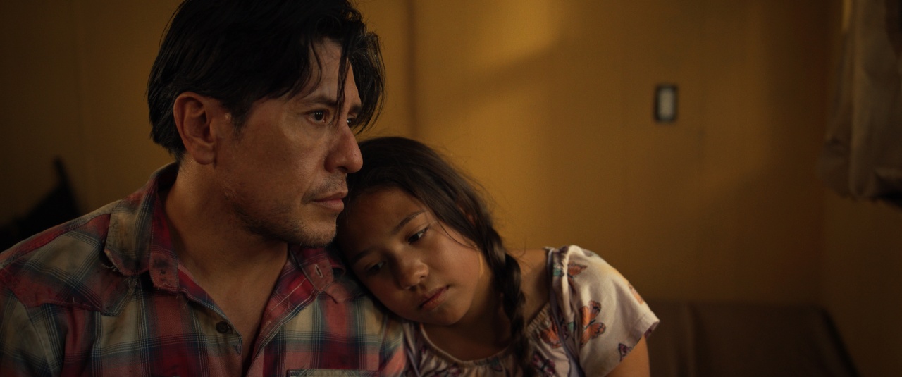 A man sits with a young girl resting her head on his shoulder; both appear thoughtful in a dimly lit room with yellow walls.