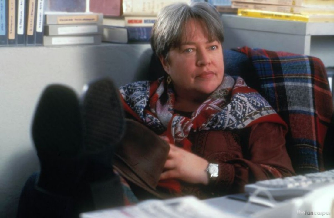 A woman with short gray hair sits at a desk with her feet up, wearing a patterned scarf and burgundy jacket, surrounded by books and office supplies.