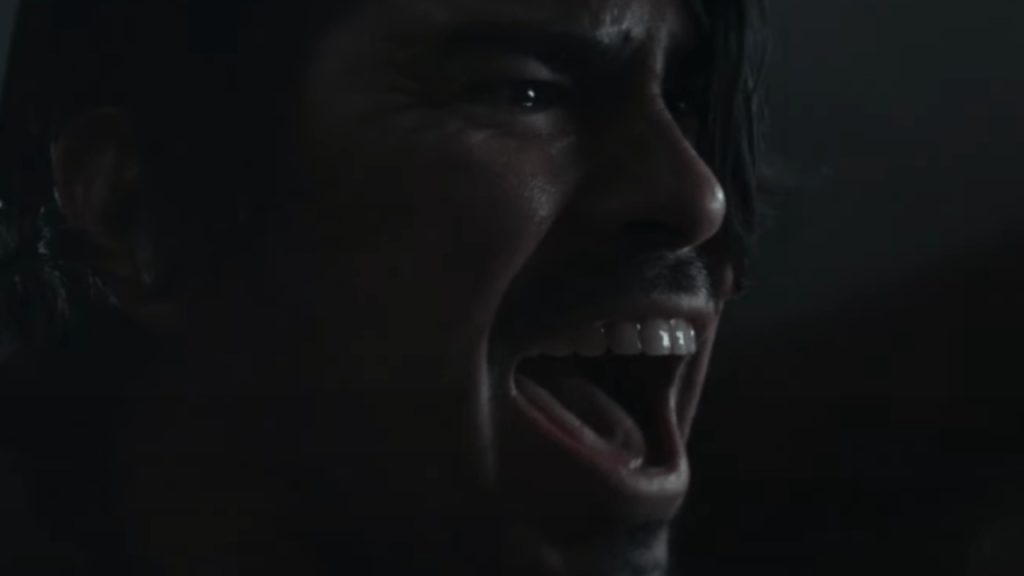 A person with dark hair is shown in close-up, mouth open wide as if shouting or screaming, with an intense and emotional facial expression.