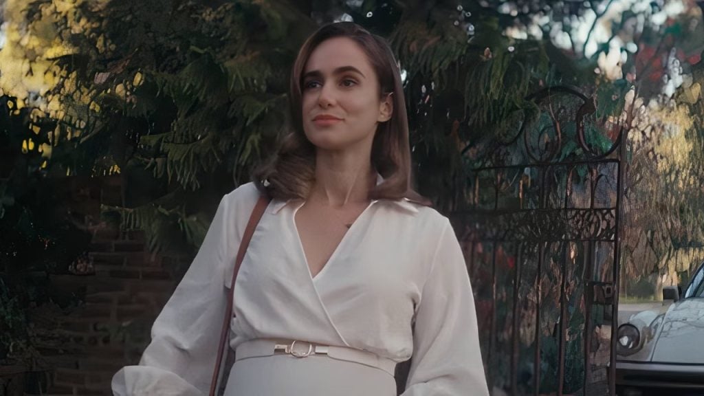A woman in a white dress stands outdoors near a metal gate and greenery, with a car partially visible in the background.