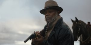 A man in a hat and coat holds a rifle outdoors, with a horse in the background under a cloudy sky.