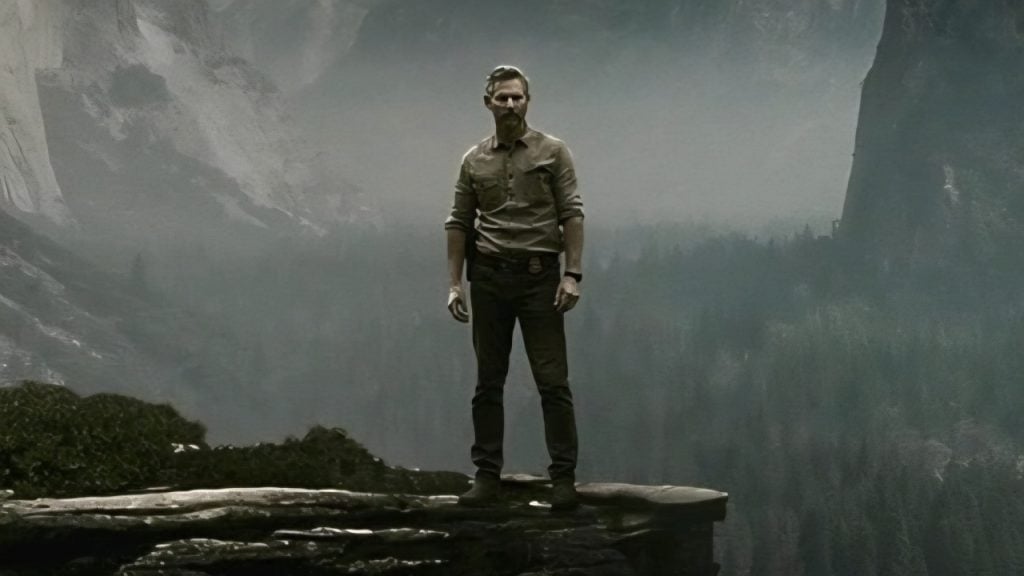 A man wearing a shirt and pants stands on the edge of a rocky cliff overlooking a misty forest and mountains in the background.