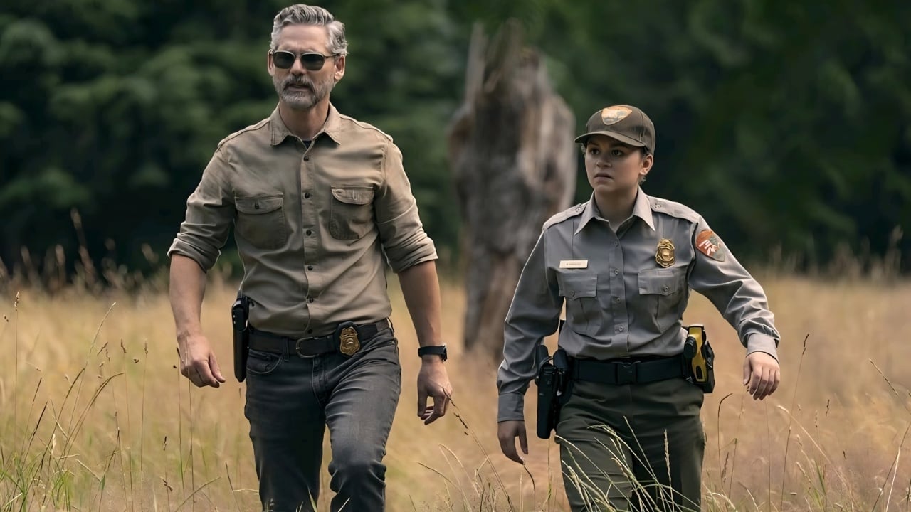 Two uniformed law enforcement officers walk through a grassy field with trees in the background.