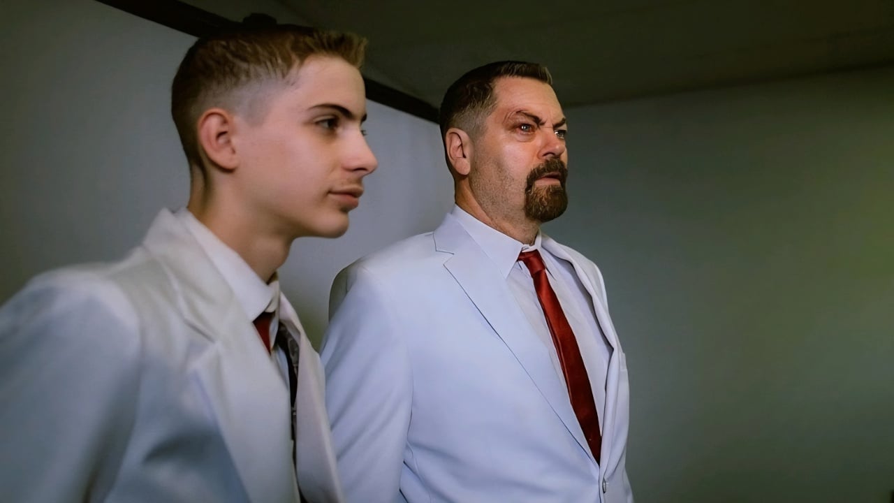 Two men in white suits and red ties stand side by side indoors, facing forward with neutral to serious expressions.