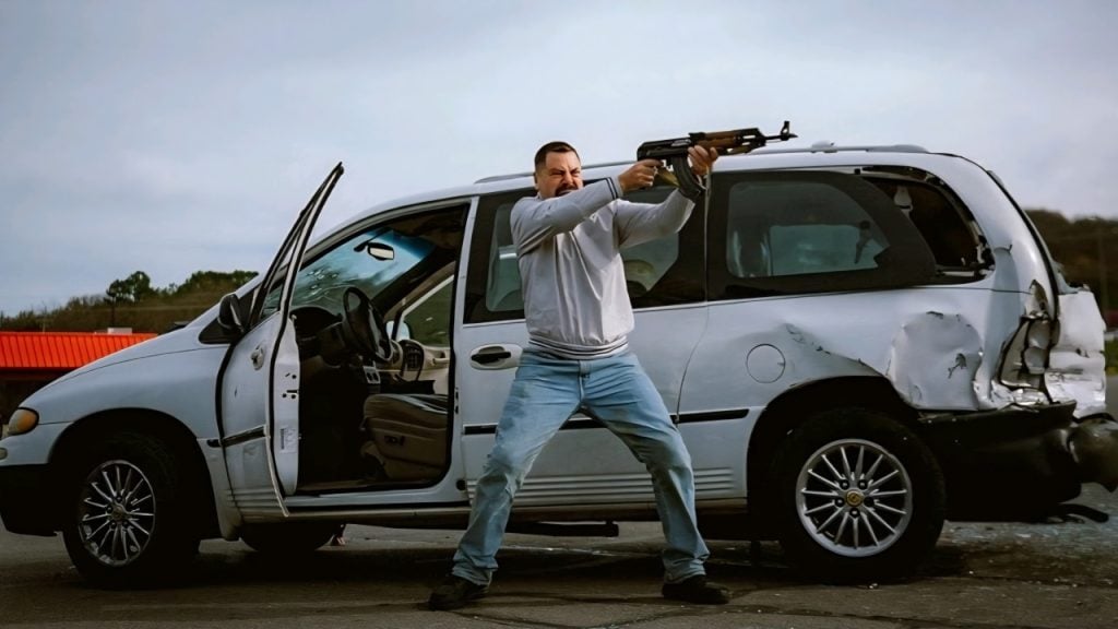 A man stands in front of a damaged minivan with the driver-side door open, holding and aiming a rifle.