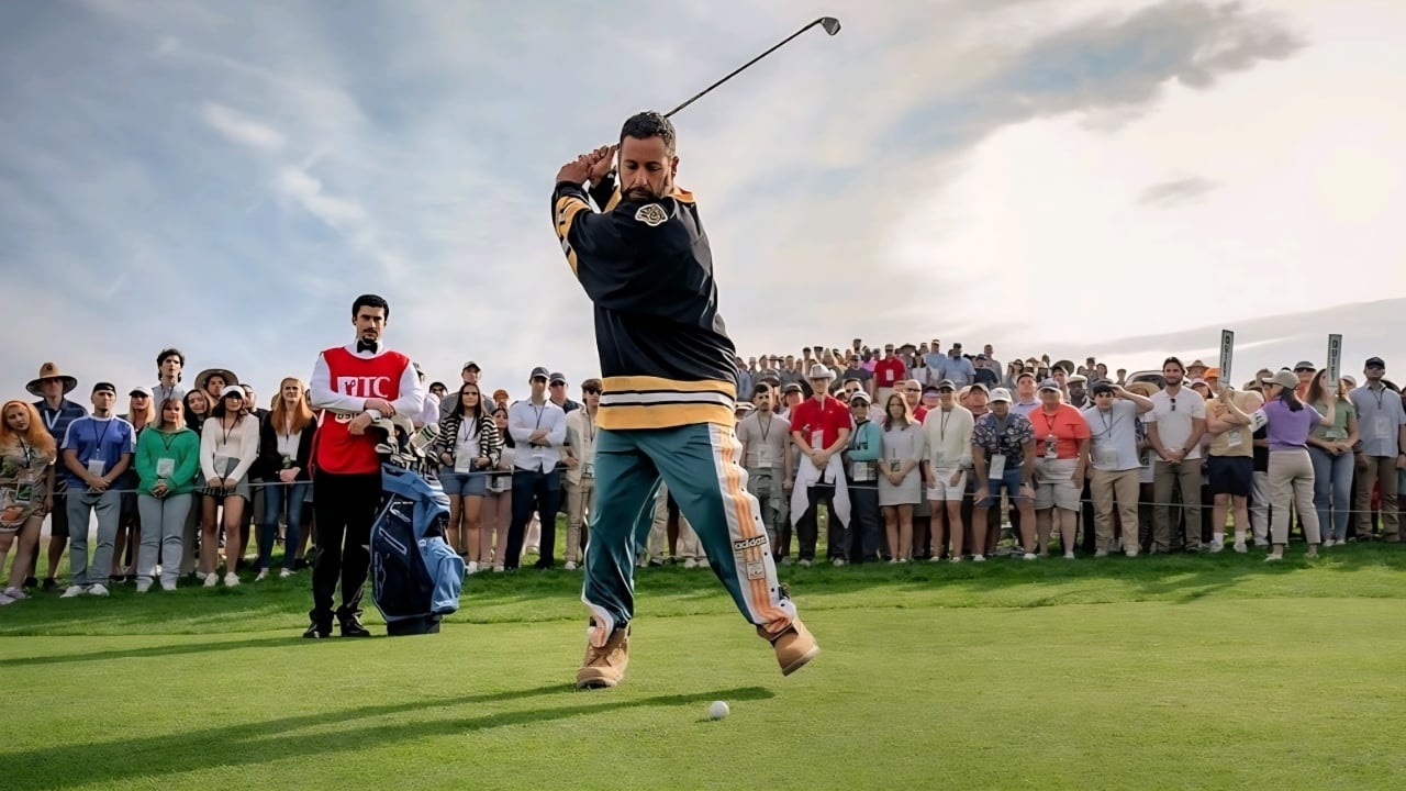 A man wearing a hockey jersey and boots swings a golf club on a course, with a crowd of spectators watching in the background.