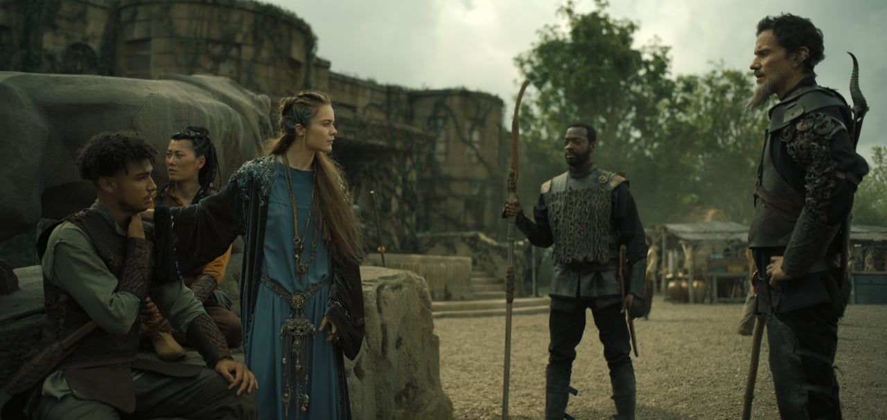 Five people in medieval-style clothing stand outdoors near a stone structure, engaged in conversation, with greenery and rustic buildings in the background.
