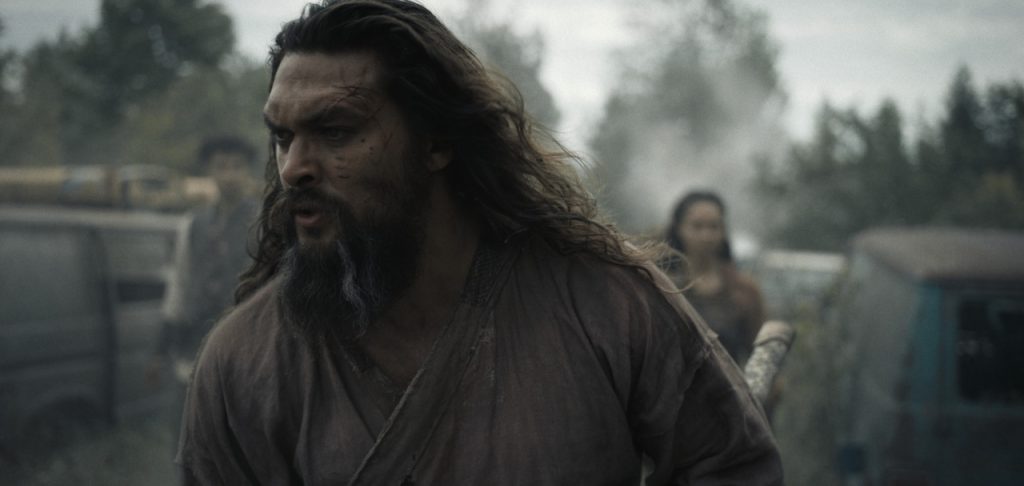 A long-haired, bearded man with face scars walks urgently outdoors, with two figures and vehicles visible in the background.