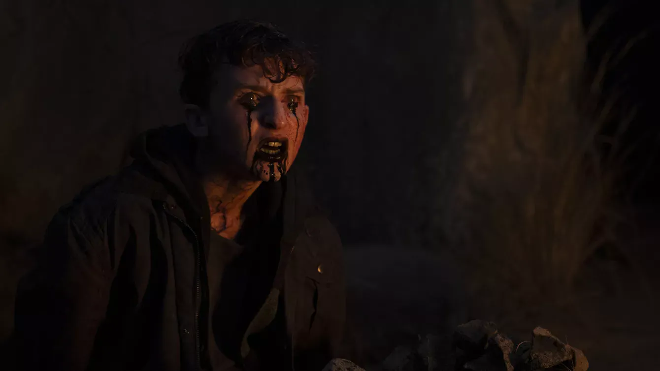 A person with blood streaming from their eyes and mouth sits in a dark, outdoor setting, appearing distressed and wearing a dark jacket.