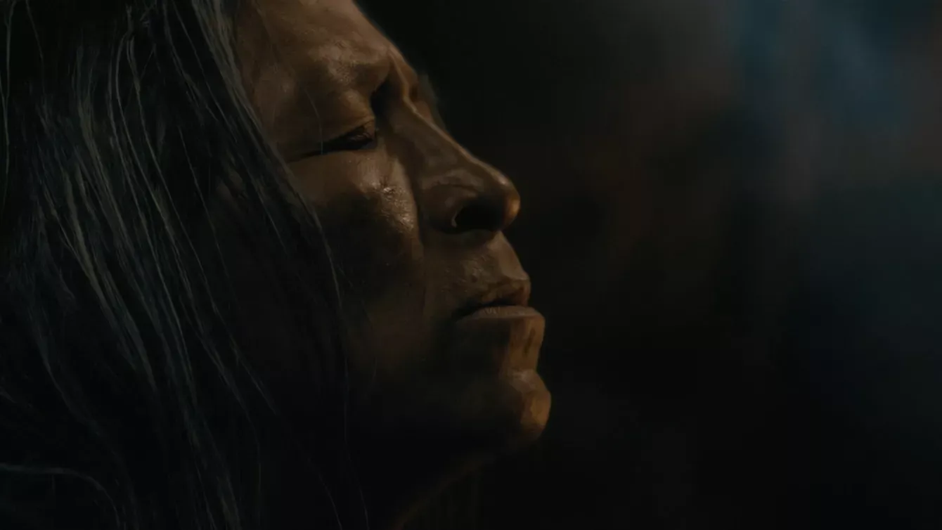Close-up side profile of a person with long dark hair and closed eyes, showing a solemn expression in low, dramatic lighting.