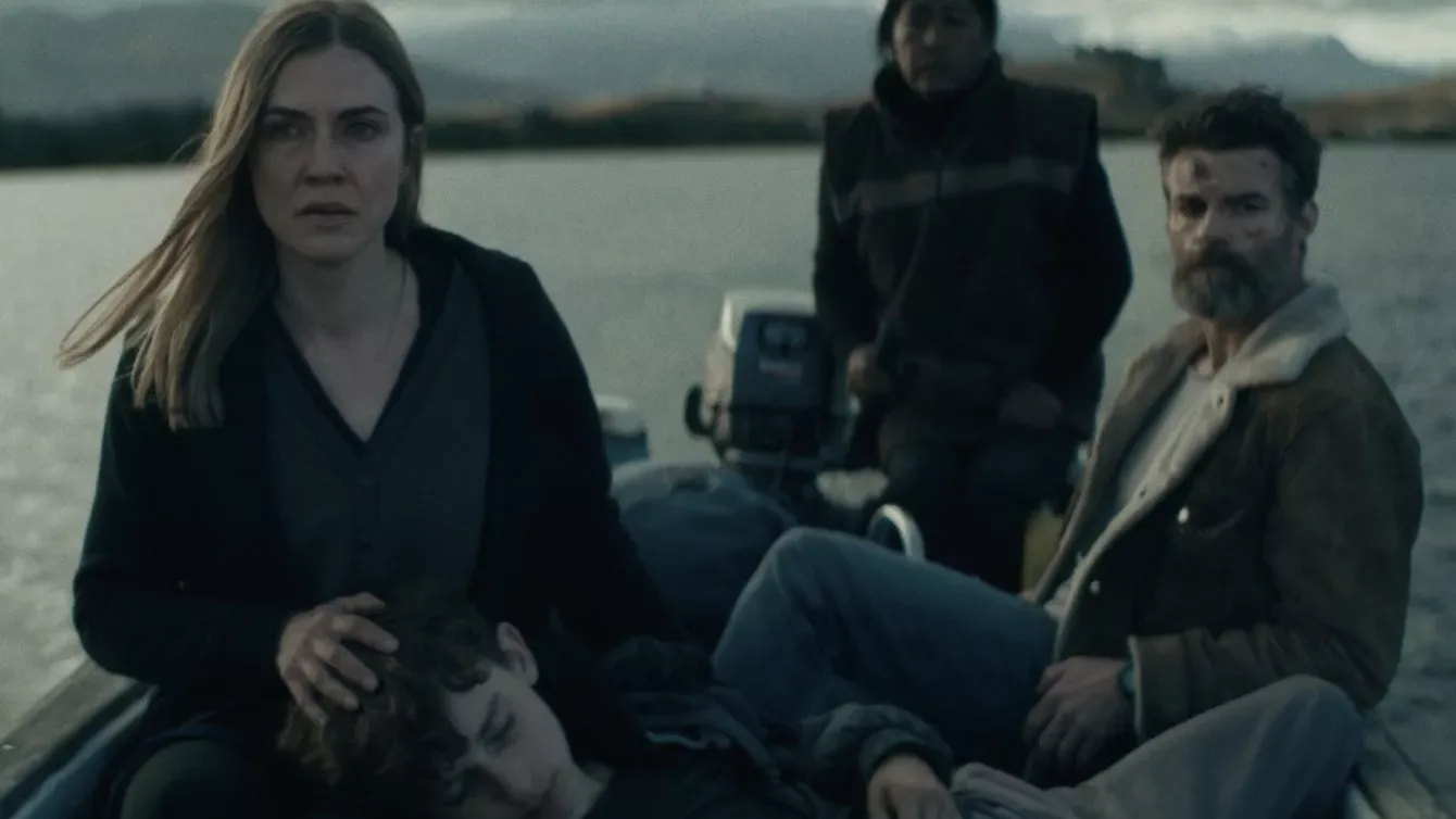 Three adults sit in a small motorboat on a lake; a woman comforts a sleeping child on her lap while the others look ahead with serious expressions.