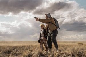 Lionsgate To Release The Taron Egerton Thriller ‘She Rides Shotgun’ On 4K UHD Blu-Ray An adult wearing a hoodie aims a handgun while standing in a dry field with a child; both look focused, with cloudy skies overhead.