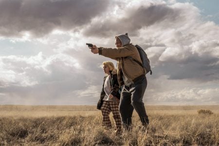 Lionsgate To Release The Taron Egerton Thriller ‘She Rides Shotgun’ On 4K UHD Blu-Ray An adult wearing a hoodie aims a handgun while standing in a dry field with a child; both look focused, with cloudy skies overhead.