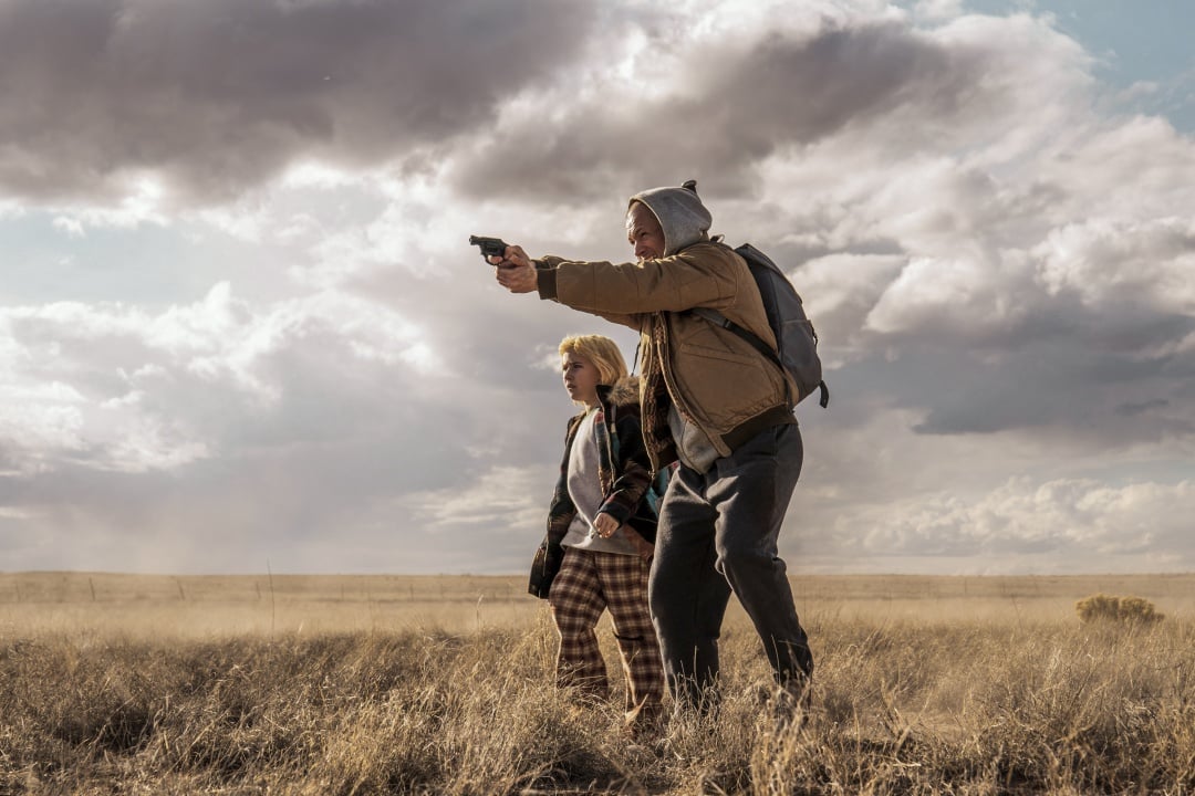 An adult wearing a hoodie aims a handgun while standing in a dry field with a child; both look focused, with cloudy skies overhead.
