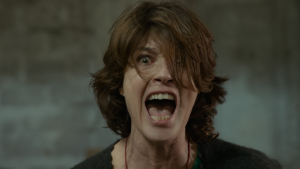 A person with medium-length brown hair is screaming with mouth wide open, eyes wide, and disheveled hair partially covering their face.