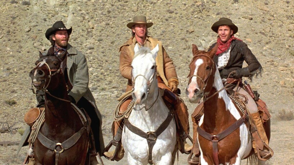 Three men wearing cowboy hats and western attire ride horses side by side in a desert landscape.