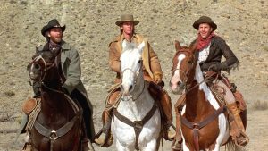 Three men wearing cowboy hats and western attire ride horses side by side in a desert landscape.