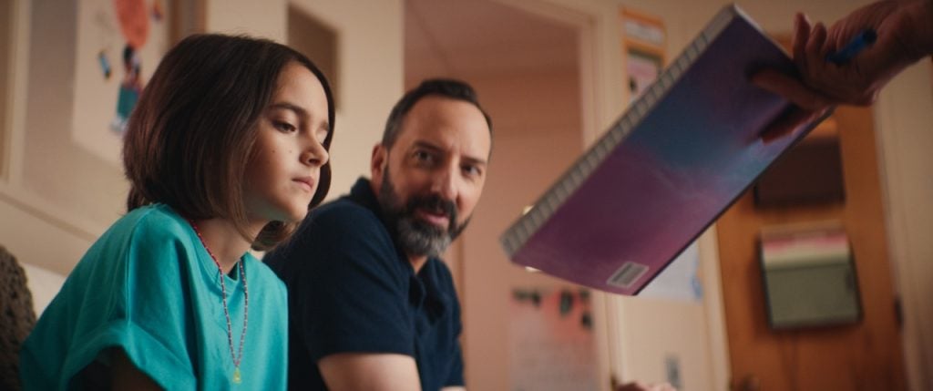 A child sits next to an adult in a room as someone hands over a spiral-bound notebook.