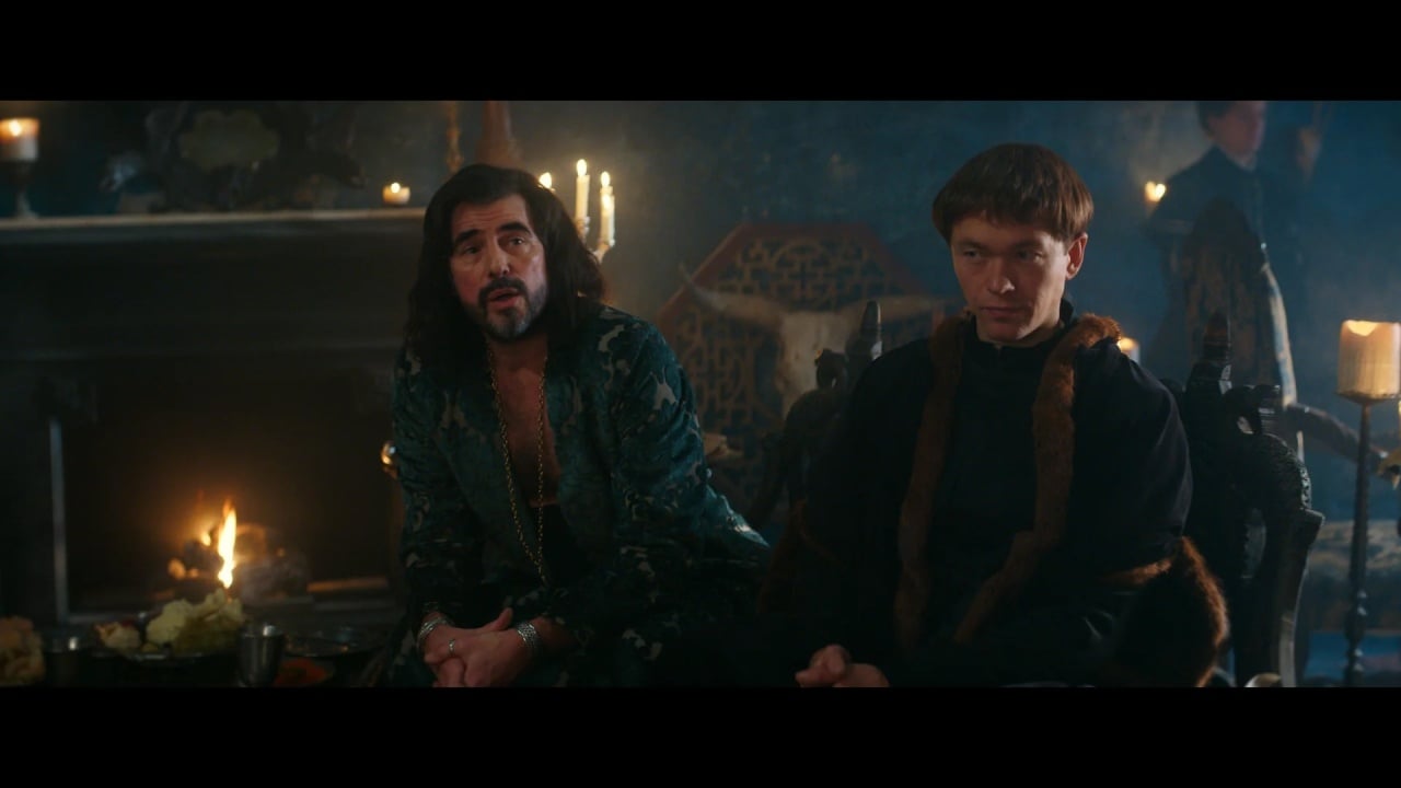 Two men in medieval-style clothing sit indoors by a lit fireplace, with candles and ornate decorations in the background.