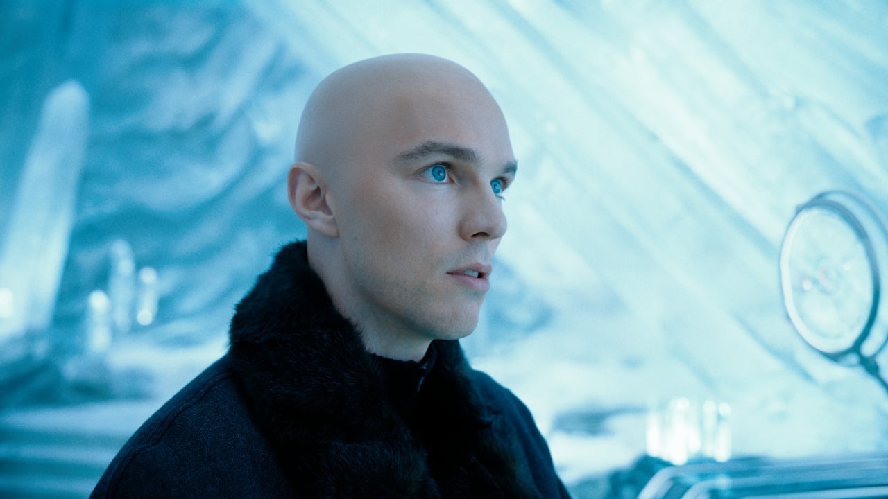 A bald person wearing a dark fur-collared coat stands in a blue, icy, futuristic environment, looking slightly to the left.