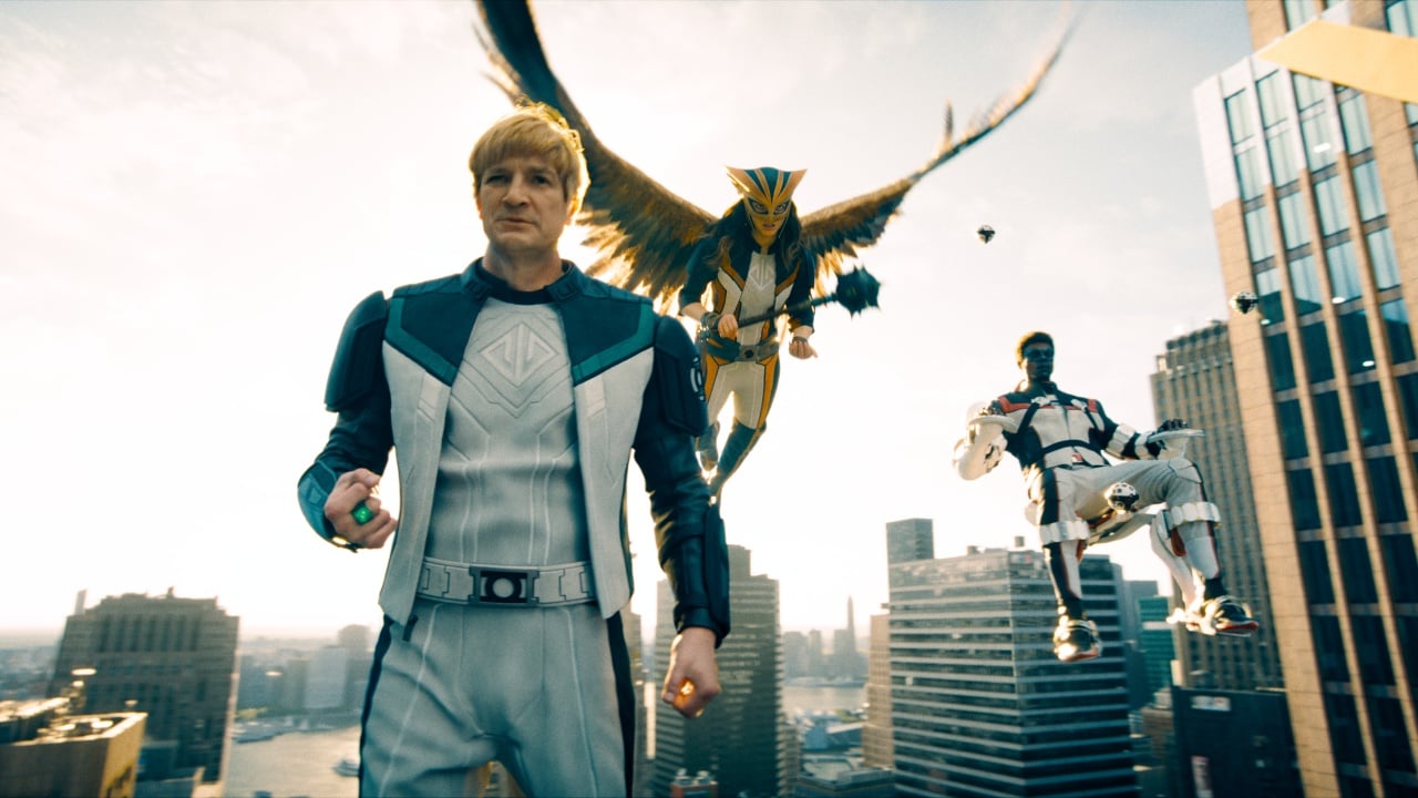 Three superheroes, one with a winged suit, one in a white armored suit, and one on a flying device, stand and hover above a city skyline with tall buildings in daylight.