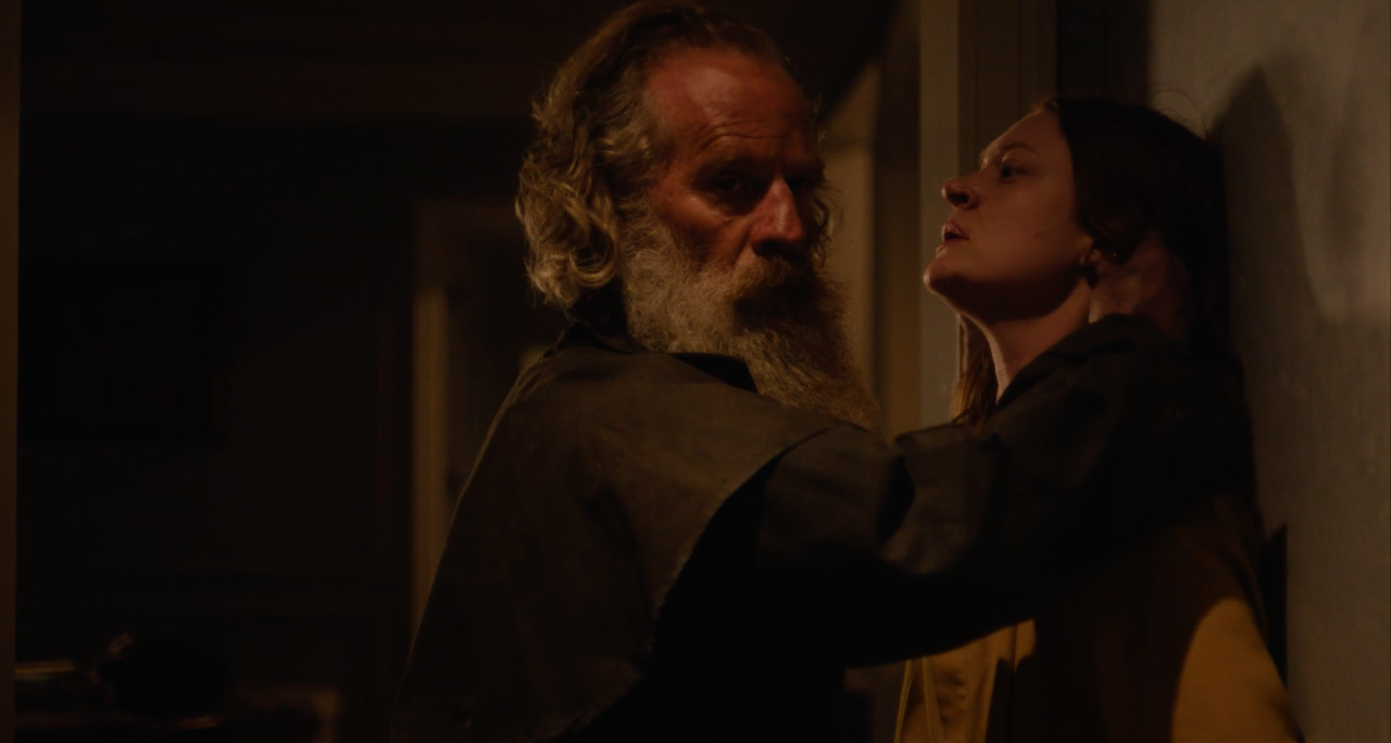 An older man with a beard forcefully holds a younger woman against a wall in a dimly lit room, both appearing tense.