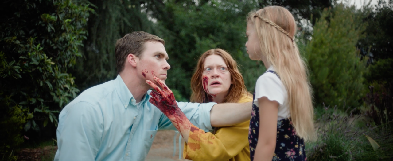 A woman with a bloody hand grabs a man’s face while looking at a young girl standing in front of them outdoors.