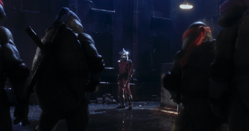 Four humanoid turtles face a figure in steel armor and a helmet in a dimly lit industrial setting.