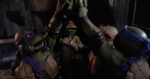 Four Teenage Mutant Ninja Turtles wearing colored masks high-five each other in a dimly lit room.