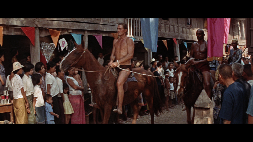 Two men wearing minimal clothing ride horses through a crowded village street during a festival, with colorful flags and onlookers lining the scene.