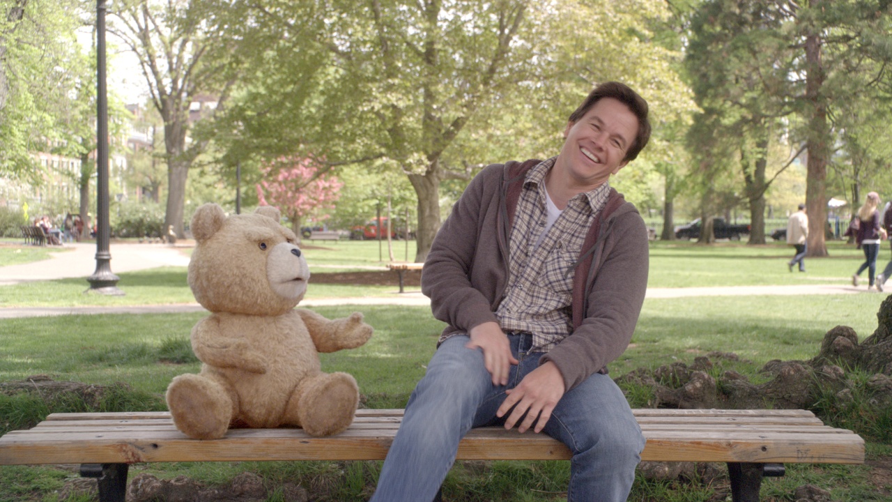 A man sits laughing on a park bench next to a teddy bear, with trees and people in the background on a sunny day.