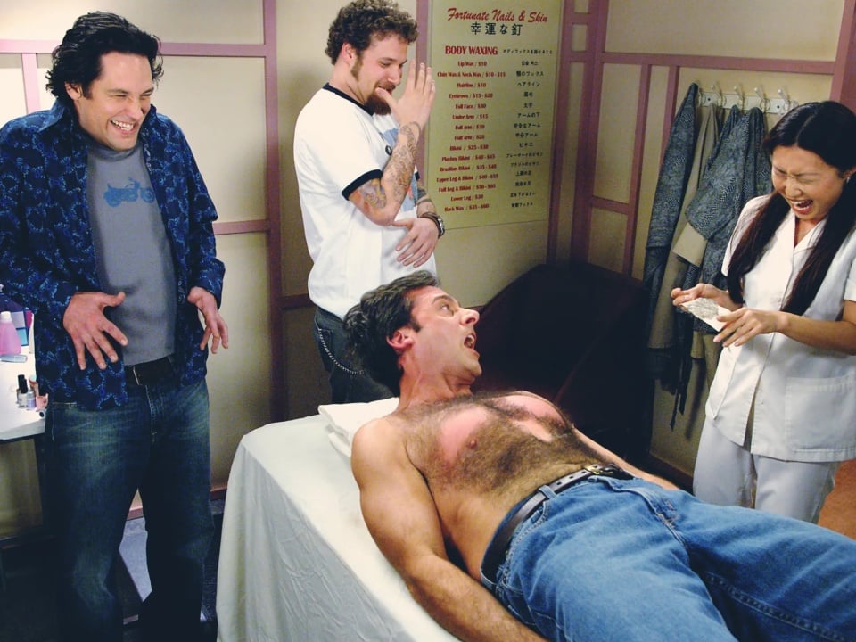 A man on a table reacts in pain while getting his chest waxed; three other people around him laugh at his reaction.