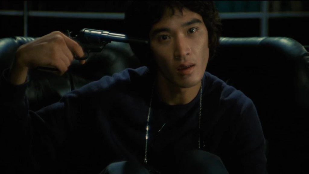 A young man sits on a dark couch, holding a revolver to his head with a serious expression, in a dimly lit room.