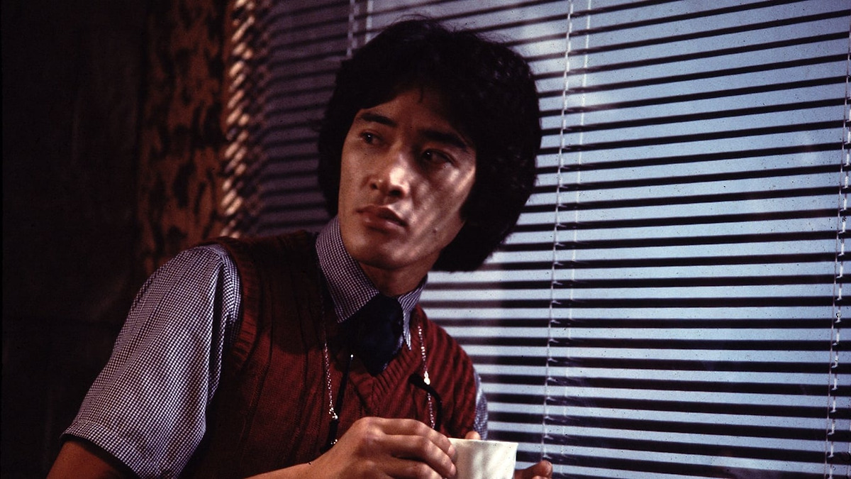 A man with dark hair wearing a checkered shirt and red vest holds a white cup while looking out through window blinds.