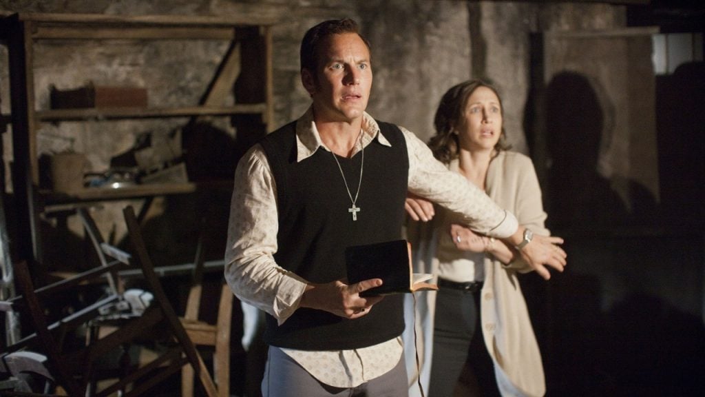 A man holding a book and wearing a cross necklace stands protectively in front of a woman in a dimly lit, cluttered room.