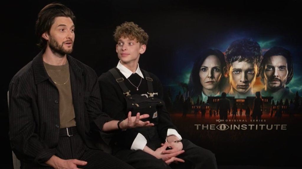 Two people sit and talk during an interview, with a poster for the show "The Institute" featuring four faces and a building in the background.