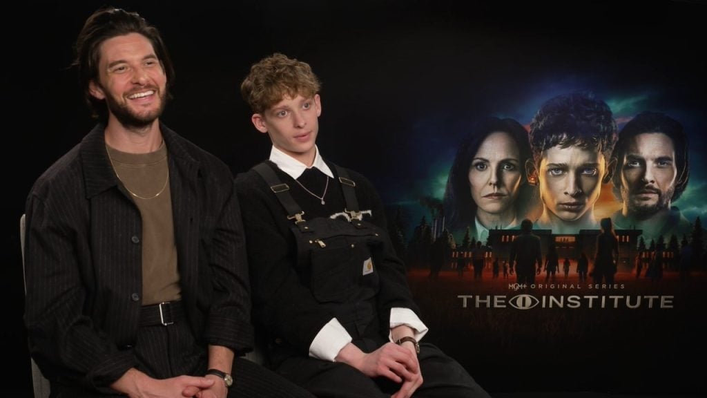 Two people are seated and smiling in front of a promotional poster for the MGM original series "The Institute," which features four serious faces and a building.